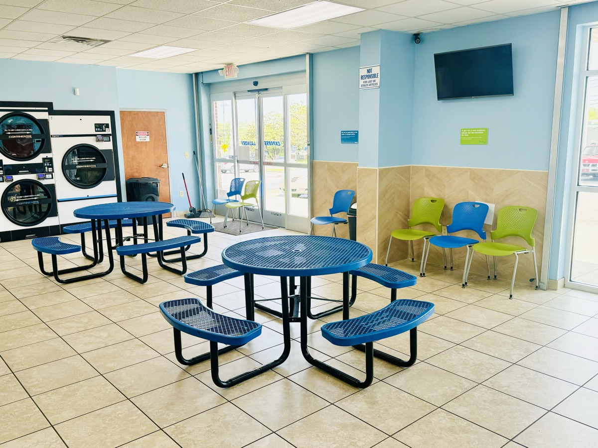 Área de espera con mesas, sillas y un televisor junto a las ventanas del frente en Express Laundry Center